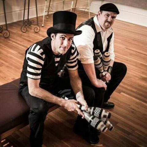 Two men in black and white outfits sitting on a stool with juggling pins.