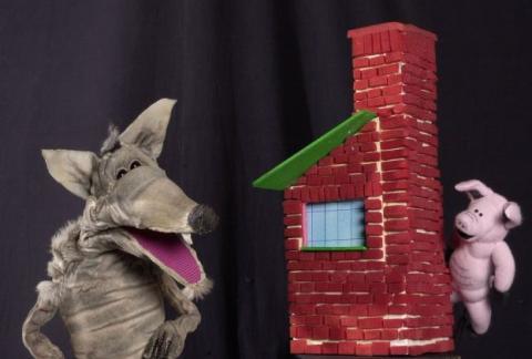 Wolf puppet standing beside a brick house and pig puppet.