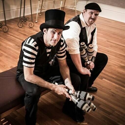 Two men in black and white outfits sitting on a stool with juggling pins.